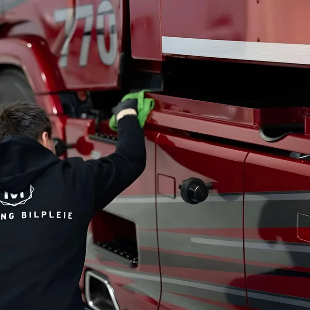 Mann i hettegenser med «Midtlyng Bilpleie» vasker rød lastebil med grønn klut.